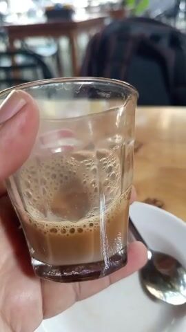 Here's a brief description of the video:

A person holds a small, clear glass filled with a light brown, bubbly liquid, likely coffee or tea with milk. The glass has a gold rim and is partially empty.  A white saucer and a silver spoon are visible on the table next to the glass. The background shows a wooden table, other chairs, and some blurred objects. The scene appears to be indoors, possibly in a cafe or restaurant.