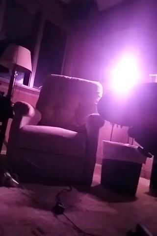The camera shows a sparsely furnished room bathed in purple light, seemingly from a light source visible through the window. In the room, a recliner chair sits prominently in the center, near a floor lamp and a small trash can.  There is a dark figure partially obscuring the view on the right side of the frame. Cords are strewn on the carpeted floor.