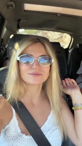 This is a first-person perspective video taken from inside a moving vehicle. The camera focuses on a blonde woman wearing sunglasses and a white top. In the background, a young girl in a pink dress with white polka dots is seated in a car seat, smiling and posing. Further back, another person is visible in the back seat. The car has a sunroof open, and the exterior environment suggests a sunny day with trees and a road visible through the windows. The overall scene appears to be a family road trip.