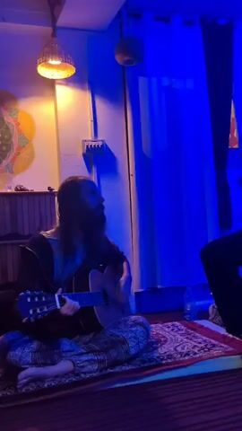 Two males are in a room with blue lighting. One male, on the left, has long hair and a beard and is sitting on the floor playing an acoustic guitar. He is wearing patterned pants and a dark shirt. The other male, on the right, is sitting on a black cajon drum and appears to be singing or talking. He is wearing a light-colored sweater with a collar and dark pants. There are some decorative items on the wall and hanging from the ceiling. A rug is on the floor between the two individuals.