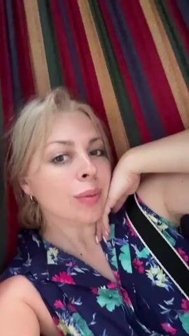 A woman with blonde hair is lying in a hammock with a colorful striped fabric. She is wearing a dark blue shirt with a floral print. Her left arm is bent and resting on her head. The camera is positioned close to her face, and she is looking directly at it.