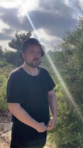 A man wearing a black bucket hat and a black t-shirt stands outdoors in a natural setting. He has a cigarette in his mouth and is holding a lighter in his right hand. The background features lush green vegetation, trees, and a cloudy sky with sunlight filtering through. The camera then pans to reveal a panoramic view of a city or town nestled among hills and greenery, with a clear blue sky and scattered clouds above. Finally, the camera focuses on a thorny bush with dense foliage.