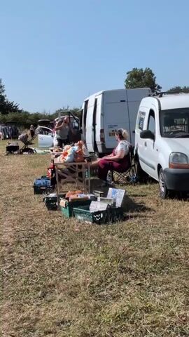 The video shows an outdoor event that appears to be a car boot sale or flea market. Several cars and vans are parked, and their trunks or rear doors are open, displaying various items for sale. People are milling around, looking at the goods, and interacting with vendors. In the foreground, a woman is sitting in a chair next to a table laden with items. A man is standing near a van in the background. The ground is covered in dry grass, and trees are visible in the distance under a clear blue sky. The scene is bathed in sunlight, indicating a warm, clear day.