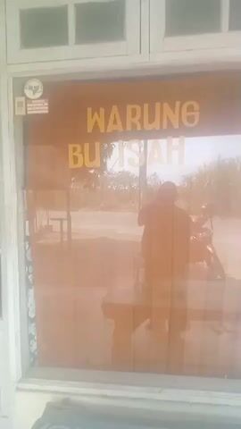 This video shows the front of a shop called WARUNG BU ISA. The shop has a wooden door and window frames. There is a QR code and a small logo on the left side of the door. The word WARUNG is written in yellow on the top part of the door, and BU ISA is written below it. The glass door reflects the scene outside, which includes a street, a motorcycle, and a person standing. The person is wearing a dark colored shirt and appears to be taking a photo or video of themselves. The environment outside seems to be rural, with vegetation and a clear sky. The video is shaky, suggesting it was recorded by hand.