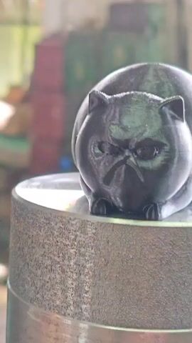 A close-up shot focuses on a black 3D-printed cat figurine with a grumpy expression. The figurine is resting on a textured, cylindrical surface, possibly a stone wheel or pedestal. In the background, out of focus, are industrial elements like pipes and machinery, with hints of green and brown colors suggesting an indoor, possibly workshop or factory setting. The lighting appears natural, coming from the right side, casting subtle shadows on the figurine. The camera slowly pans to the right, revealing more of the cat's body and the surrounding environment.