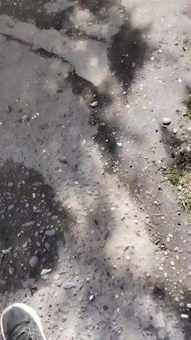 The camera pans over a patch of wet dirt, small rocks, and the shadow of a tree, before focusing on a narrow, shallow stream. The stream flows from under a concrete culvert with a grate. Grass grows along the edges of the stream and a small tree trunk is also visible in the background. The water in the stream is clear enough to see the dark stones at the bottom.
