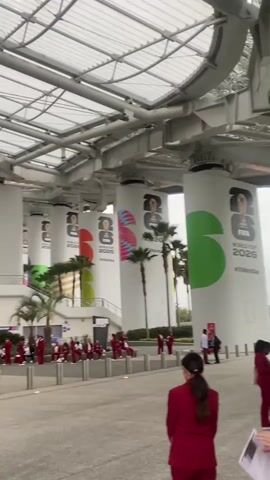 The video shows a group of people, mostly wearing red suits, gathered under a large, modern structure with a translucent roof. The structure appears to be an outdoor walkway or entrance area of a stadium or large venue, supported by massive white cylindrical pillars.

Each pillar is decorated with large graphics. The most prominent graphics are for the "FIFA World Cup 2026," featuring the FIFA logo and the year. Some pillars also display abstract colorful designs and images of soccer players. Palm trees are visible behind the pillars, suggesting a warm climate.

The people in the foreground are facing away from the camera, while a larger group is seated or standing in rows further back, also facing the same direction. Some individuals in red suits appear to be staff or performers. A few other people in business attire are also visible, possibly officials or guests. The overall atmosphere seems to be one of anticipation or preparation for an event. The lighting suggests it is daytime, with overcast weather. The presence of FIFA branding clearly indicates that this scene is related to the 2026 FIFA World Cup, which is scheduled to be hosted in the United States, Canada, and Mexico. The location context of Inglewood, USA, further points to a venue in that region preparing for the tournament.