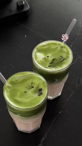 Here is a detailed description of the video:

The video features a close-up, overhead shot of two glasses filled with a layered beverage, likely a matcha latte. Each glass has a light-colored base, probably milk, topped with a vibrant green layer of matcha. Ice cubes are visible within the green layer.

A hand with manicured nails enters the frame and uses a clear plastic straw, adorned with a small flower decoration, to stir the contents of each glass. The action mixes the matcha and milk layers. The background is a dark, possibly black, countertop with a blurred object in the upper left corner. The lighting is bright, highlighting the colors and textures of the drinks.