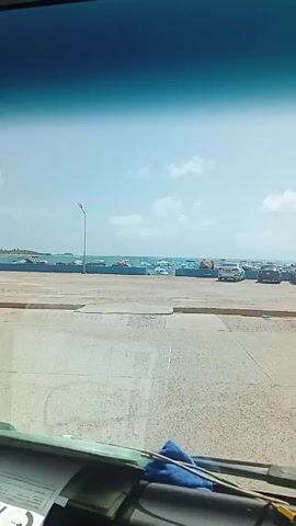 The camera records a view from inside a car. Looking through the windshield, a concrete area is visible, leading to a shoreline with boats and a line of parked cars. The sky is blue with scattered clouds. A sticker labeled 'COBRETAG' is visible on the rear view mirror.