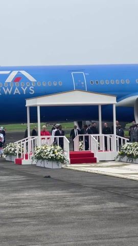The video captures a formal outdoor ceremony taking place on an airport tarmac in Malabo, Equatorial Guinea. A large blue airplane, clearly marked with "ITA AIRWAYS" and partial registration numbers, dominates the background.

In the foreground, a white ceremonial platform with red carpeted steps is set up, adorned with numerous white and yellow floral arrangements. A central figure, dressed in distinctive red ecclesiastical robes, stands on the platform, facing towards the left. He is surrounded by several other men in dark suits and various military and official uniforms, suggesting a high-profile dignitary's arrival or departure. To the far left, a woman in colorful traditional African attire walks along the tarmac.

The sky is overcast, indicating a daytime event under cloudy conditions. The overall atmosphere is formal and respectful, with ceremonial music audible in the background.