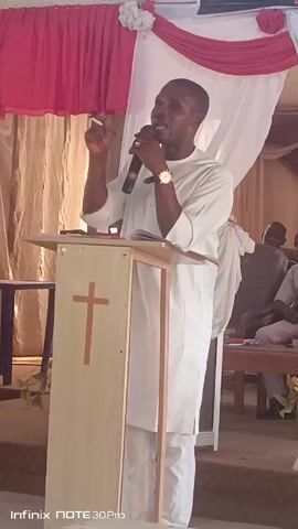 A man in white stands behind a light-colored podium, speaking into a microphone. The podium has a simple cross design on its front. The man is wearing a watch.  Behind the podium, there is draped fabric in white and red. Several seated people are visible in the background. The man gestures with a pen in his left hand while he speaks. A small table is off to the left of the podium. The words "Infinix NOTE 30 Pro" are at the bottom of the frame.