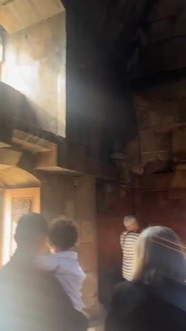 The video captures an indoor scene within an old stone building, likely a church or monastery, characterized by rough-hewn stone walls, carved details, and arched doorways. Sunlight streams through a high window, illuminating dust motes in the air. A group of people are present, including several adults and one child held by an adult. A man in a white t-shirt carries a silver tray with items on it, while another man next to him holds a stick with multiple lit candles. Other individuals stand observing or slowly move through the space. The camera pans around, revealing more of the stone architecture, including relief carvings on the walls and a large arched entrance with steps.