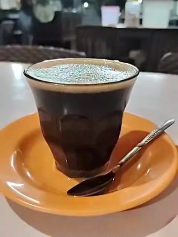 A cup of dark liquid, likely coffee, rests on a small orange saucer on a white table. A spoon rests beside the cup on the saucer. The cup is faceted and clear, allowing the dark liquid to be seen. Bubbles cover the surface of the liquid. The background shows a blurry interior of what may be a cafe or restaurant. There are wicker-style chairs and other tables visible. The lighting suggests it is indoors and possibly daytime.