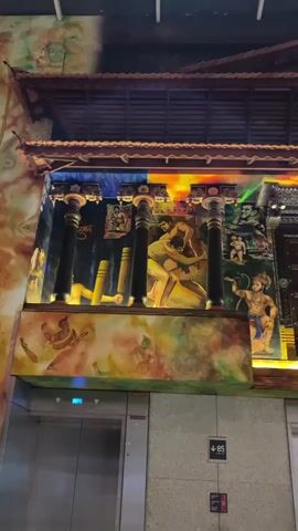 A vertical video pans across a mural painted on a building facade. The mural features several distinct scenes depicting traditional Indian themes. In the center, two muscular men are engaged in a wrestling match, rendered in a realistic style. To the left of the wrestlers, a scene with stylized yellow cylindrical objects suggests a sport or activity. To the right, a depiction of Hanuman, a Hindu deity, is prominent, along with smaller vignettes of other figures. The mural is illuminated by colored lights that create a vibrant, multi-hued effect. The top of the structure has a dark, tiled roof with ornate wooden trim. Below the mural, an elevator door with a blue light is visible. The overall aesthetic suggests a cultural or artistic display, possibly at a public venue.