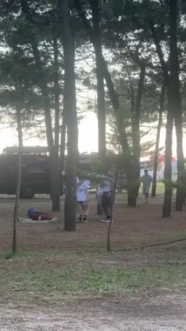 The video shows a group of people in a wooded area. There are tall trees casting shadows on the ground. A dark-colored van is parked nearby. People are standing and talking, while one person walks into the scene from the right. Two girls walk through the scene from left to right. In the background, someone shouts in a non-English language. A red bag lies on the grass. The sun appears to be setting or rising.