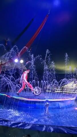 A person in a red costume rides a unicycle in a circle in the middle of a fountain. Water sprays up around the unicyclist. The background is dark blue with spotlights and red fabric hanging from above.