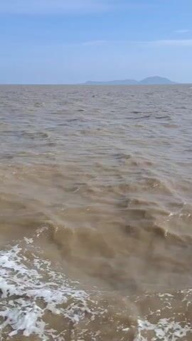 The video shows a choppy, muddy body of water with whitecaps breaking near the bottom of the frame. The water stretches to the horizon where a faint landmass with hills is visible under a clear blue sky. The camera appears to be positioned on a boat or a pier, looking out over the water.