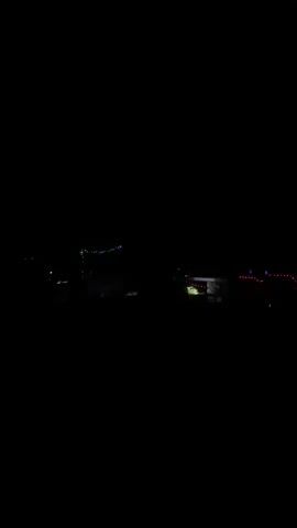 A shaky, dark video captures a nighttime scene with distant, faint lights. In the center, a structure with colorful string lights is visible, possibly a building or a tent. To the right, another illuminated structure with red lights is present, appearing to be further away. The overall environment is dark, suggesting a rural or outdoor setting at night. The camera movement is unsteady, making it difficult to discern specific details or activities.