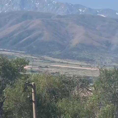 The video shows a landscape with mountains in the background and trees in the foreground. The mountains appear to be rugged and have some snow on their peaks. The trees are green and leafy, and they seem to be in a natural setting. The sky is clear and blue. The camera is panning slowly to the right.