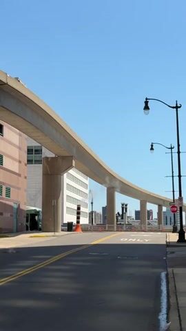A bright, clear day with a deep blue sky. A concrete monorail track curves from left to right across the frame, supported by thick, grey pillars. To the left, a multi-story building with brown and beige panels and green ventilation grates stands beneath the monorail. A green bus is partially visible entering an opening in the building. In front of the building, an orange traffic cone is placed on the ground. Beyond the monorail, the city skyline is visible with several tall buildings, and two lampposts stand on the right side of the frame. A white SUV is seen driving under the monorail. The road in the foreground has two yellow lines down the center and a white line on the right edge.