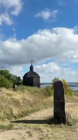 The video shows a landscape view from a hilltop. In the foreground is a small, wooden chapel with a cross on top, and a stone monument with inscriptions. The midground consists of a grassy slope and some scattered trees. In the background, a wide river or lake stretches out to the horizon, with green islands and trees dotting the water. The sky is blue with scattered white clouds. The camera pans slightly to the right, revealing more of the river and surrounding landscape. The overall impression is one of a peaceful, rural setting.