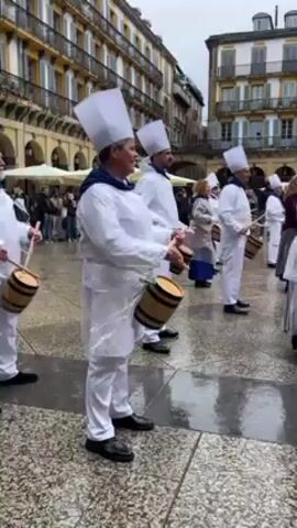 On an overcast day, a group of people in matching white outfits and chef's hats perform in a wet, European city square. The majority of the group is drumming on small barrels. A single individual in the same attire appears to direct the group from the front. A crowd of mixed ages and dress watch in the background near a building.