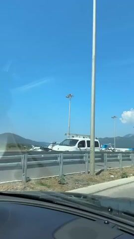The video is a view from a car on a sunny day, looking out at an airport tarmac. Several small to medium-sized airplanes are parked on the tarmac. Some of the planes are white with red and blue accents, while others are predominantly blue. There are tall light poles on the tarmac. In the background, there are hills or mountains and a clear blue sky. The video pans slightly to the right, showing more of the parked aircraft.