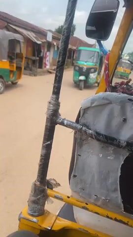 The video shows a view from inside a vehicle, likely a tricycle or tuk-tuk, driving on a dirt road in a rural or semi-urban area. The road is lined with buildings, some appearing to be shops or homes with corrugated metal roofs. There are a few people visible on the side of the road, including a woman walking away from the camera and a man sitting on a step. The vehicle appears to be in motion, as indicated by the blurred background and the perspective. The interior of the vehicle shows some worn-out upholstery and reinforced metal bars, suggesting it may be used for public transportation. The sky is partly cloudy.