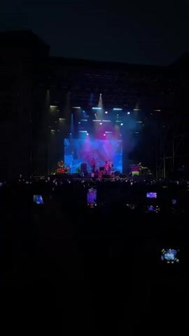 The video captures a live outdoor concert at night. The stage is illuminated by bright blue, green, and pink lights, with abstract digital graphics displayed on a large screen behind the performers. Four musicians are visible on stage. On the right, a man wearing a cap and a light-colored jacket is singing and dancing, gesturing with his hands. To his left, another person stands near a microphone, playing an instrument. Further left, two more individuals are also playing instruments. A crowd of spectators is visible in the foreground, many holding up smartphones to record the performance. The atmosphere is dark, with the stage lights being the primary illumination source.