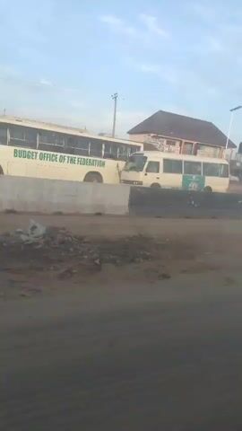This video shows a street scene in what appears to be an African country. In the foreground, there is a dirt road with tire tracks. In the middle ground, there is a concrete barrier separating the road from a street with several buildings. A white bus with green lettering is parked on the street. In the background, there are several buildings, including a two-story building with a sign that reads "SECRET OFFICE OF THE." There are also several cars and motorcycles on the street. The sky is a clear blue with a few clouds. The video appears to have been filmed from a moving vehicle.