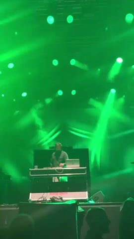 The video shows a DJ performing on a stage with green lighting and fog. The DJ is wearing headphones and is standing behind a console with a laptop and DJ equipment. The audience is visible in the foreground, with their silhouettes illuminated by the stage lights. The Red Bull logo is visible on the DJ booth.