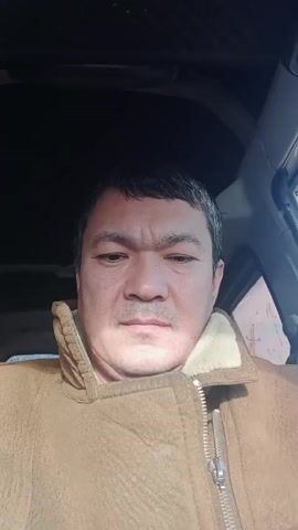 A close-up shot depicts a middle-aged man with short, dark hair wearing a brown jacket with a light-colored, furry collar. He is positioned inside a vehicle, possibly the driver's seat, as suggested by the dashboard and side window visible in the background. His expression appears neutral, and he is looking towards the camera. The lighting suggests it is daytime.