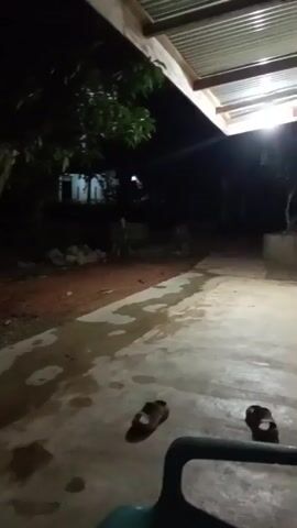 The video is a shaky, low-light recording taken at night. It appears to be an outdoor scene, possibly a rural or semi-rural area. The ground is a mixture of dirt and a concrete-like surface, with puddles of water visible on the concrete. In the foreground, a pair of brown sandals lies on the concrete. A dark-colored object, possibly a chair, is partially visible in the bottom right corner. Further into the scene, trees and some rudimentary structures or buildings are dimly lit by artificial lights. The overall impression is a very dark, possibly damp, outdoor environment at night, with minimal activity or clear detail visible.