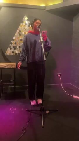 A young woman wearing headphones and a blue hoodie with a red accent over a red shirt stands in a recording studio in front of a microphone. She holds a phone in her right hand and appears to be singing or speaking into the microphone. She is wearing black pants and white sneakers. The studio has dark gray walls, a dark floor with pink lighting, and a decorative circular wall hanging with a geometric pattern of circular elements. A microphone stand is positioned in front of the woman, and a small table is visible to her left. An electrical outlet and cords are visible on the wall behind her.