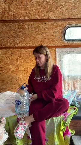 A young woman with blonde hair, wearing a maroon tracksuit and floral clogs, sits cross-legged on a bed inside a room with wood-paneled walls. She is holding a large, clear plastic bottle of water and drinking from it. Pillows with colorful patterns are visible on the bed.
