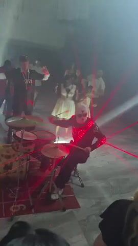 A group of people are in a dimly lit room with red and blue lights. A drummer in a white helmet and a black outfit with red lights is playing a drum set. Red laser beams are visible in the scene. Some people are dancing in the background. The drummer's helmet has a reflective visor and the outfit has illuminated accents on the chest and arms. The drummer is seated and actively playing the drums. There are other people standing and observing.