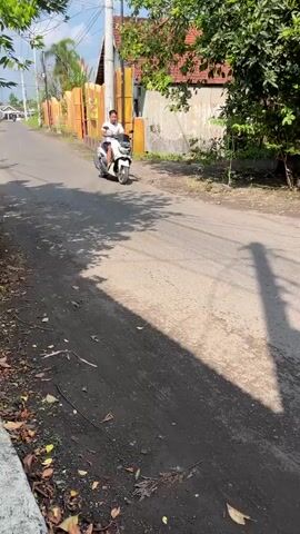 A man wearing a white t-shirt and dark shorts is riding a white scooter down a paved road. The scooter is a Yamaha NMAX, identifiable by its design. The man is looking to his left. The road is lined with trees and vegetation on the right side, and a building with yellow accents is visible on the left. The sun is shining, casting long shadows. The overall scene appears to be in a rural or suburban area.