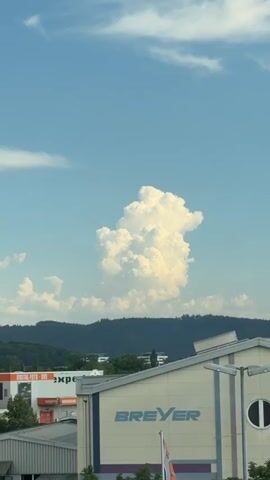 This media file captures a bright, daytime scene with a prominent cumulus cloud formation dominating the sky. The cloud is voluminous and textured, with areas of bright white where sunlight hits it directly and softer, gray shadows within its depths. It appears to be a warm, sunny day with scattered smaller clouds in the clear blue sky.

In the foreground, the top of a building is visible, featuring a light-colored facade with horizontal lines and industrial-style lighting fixtures. The name "BREYER" is clearly emblazoned in blue capital letters on this building. To the left, another building or structure displays prominent signage. One sign reads "DIGITAL-FOTO-DVD" followed by "exper".

The middle ground shows a dark, forested hillside, likely a mountain range, against which the clouds are silhouetted. The trees are dense and dark green, indicating a lush natural environment. Above the treeline, a few modern buildings with light-colored walls and dark roofs are partially visible, suggesting an urban or suburban area nestled near the natural landscape.

The overall impression is one of a pleasant, possibly late afternoon, day in a location that blends natural scenery with commercial development. The focus of the image is the dramatic cloud formation, which adds a dynamic element to the otherwise calm and sunny weather. There are no people or animals visible in the frame. The location is identified as Singen (Hohentwiel), Germany.