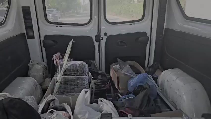 The video shows the interior of a white van with its rear doors open. The van is filled with various items, including bags, boxes, and what appears to be a dismantled metal structure wrapped in plastic. The items are jumbled together, suggesting a transfer or storage of goods. The environment outside the van, visible through the open doors, appears to be a street with parked cars and a building in the background. The lighting suggests it is daytime.