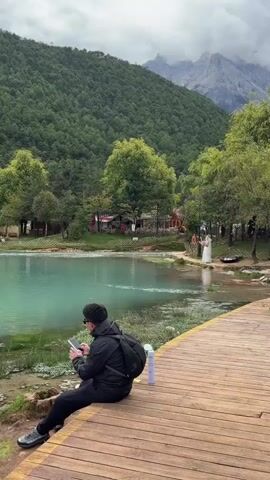 A person in a black jacket and black pants is sitting on a wooden boardwalk, looking at a smartphone. They have a black backpack on their shoulders and a white water bottle next to them. The boardwalk is next to a turquoise lake. In the background, there is a lush green forest covering a mountain, with a glimpse of snow-capped mountains in the distance. Several trees are scattered around the lake, and there are some small buildings and people visible on the opposite side of the lake. The sky is overcast.