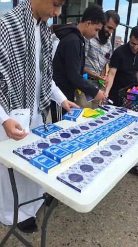 Several people are standing around a white table filled with copies of the Quran. They appear to be packaging the books with other items, possibly for distribution. One person is wearing a traditional checkered scarf, and another is wearing a black hooded sweatshirt. The background shows a building with glass doors and windows. The scene appears to be outdoors, possibly in front of a religious or community center.
