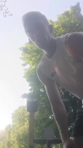 A man in a white tank top and black shorts stands outdoors next to a weight machine with two black weights. He is facing away from the camera, looking up at the sky and trees. The sun is shining brightly, creating a lens flare in the upper left corner of the frame. The man is performing an exercise.
