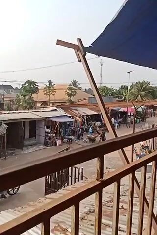 The video shows an outdoor market scene from an elevated viewpoint. The market consists of small stalls with corrugated iron roofs and various goods displayed for sale, including clothing and bags. Several people are visible in the market area, including vendors and potential customers. A man is pushing a handcart loaded with goods, and motorcyclists are driving through the market area. In the background, there are buildings and trees. A tall telecommunications tower is visible in the distance. The view is partially framed by wooden support structures and a blue awning. The overall impression is of a bustling, active marketplace.