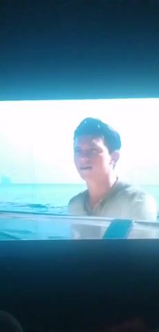 The video shows a young man in a boat on the water. He is smiling and looking to his right. The water is blue and there are some islands in the background. Then the video shows a shot from inside a cave looking out at the water. There is a small boat in the distance. The cave is dark and rocky. The water is calm and there is a bright light coming from outside the cave.