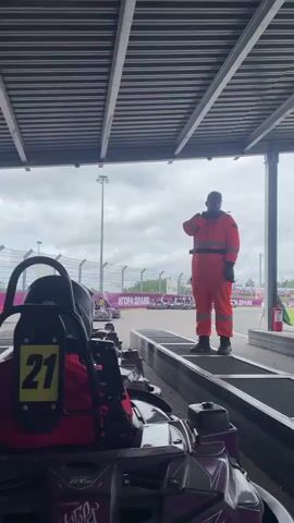 The video begins with a view of go-karts parked in a covered area. A person in an orange jumpsuit is standing on a platform, facing away from the camera. The go-karts are lined up, and some have numbers visible, such as "21". The background shows a race track with a checkered flag pattern and barriers. The sky is overcast. The person in the orange jumpsuit turns and walks away from the camera towards the race track. The camera then pans slightly to show more of the go-karts and the track. The video ends with a shot of the person in the orange jumpsuit walking away.