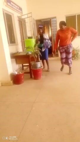 Two women are in a hallway. One woman wears an orange long-sleeved shirt and patterned capri pants, walking towards the camera and then out of view. The second woman stands in front of a desk, watering a potted plant with a green bowl. She wears a dark top, a blue skirt, a shawl over her shoulders, and sandals. There are two red buckets on the floor. Above the desk is a sign that reads "Department of Social Science Education." There is a door to a room that says "Exam Office." The floor is tiled and there are windows on either side of the doorway. A TECNO POVA watermark is in the lower left corner.