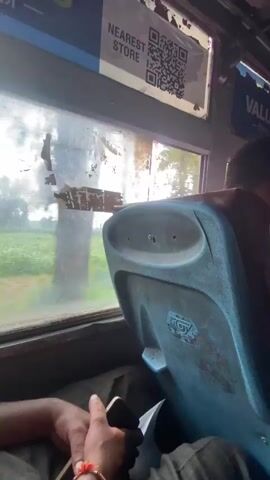 The video provides a first-person perspective from inside a moving vehicle, likely a bus. The camera is pointed towards the right side, showing a window with a blurred, green, outdoor landscape passing by. A predominantly blue, worn seatback is in the foreground, obscuring part of the view. Above the window, a sign with a QR code and the text "NEAREST STORE" is visible, along with other indecipherable text. A person's hand, wearing an orange bracelet, is holding a smartphone in the lower left corner of the frame. Further back in the bus, other passengers and seatbacks are visible, with sunlight streaming in from the windows, creating a bright, slightly hazy atmosphere. The overall impression is that of travel in a crowded public transportation vehicle.