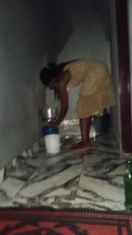 A woman with dark skin and long dark hair is stirring something in a pot. She is wearing a yellow dress with a brown and white floral pattern. She is standing barefoot on a marble-patterned tile floor. The pot is on a small blue gas stove, which is sitting on top of a white bucket. There is a red rug in the lower foreground of the frame, and a green glass bottle near the bottom right corner. The room appears to be a small kitchen or storage area. The walls are light gray. There is a black bucket in the background. The lighting is dim.