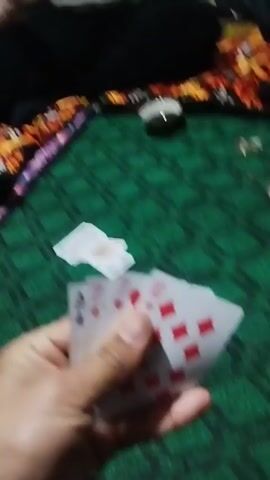 Here is a brief description of the video:
The video features two people sitting on a green patterned rug, playing cards.  The primary focus is on one of the individuals who is looking directly at the camera and speaking. The camera is held at eye level and occasionally shows the other person, cards scattered on the rug, and a small cup. The individual speaking is wearing a light brown zippered sweater.