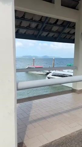 A slow pan right reveals a view from a covered structure looking out over a body of water towards a pier. In the foreground, there is a tiled floor and a white railing. Beyond the railing, the blue water is visible with a small white boat docked. The pier extends into the water on the left, with several buildings and structures visible on it. The sky is mostly blue with some clouds.